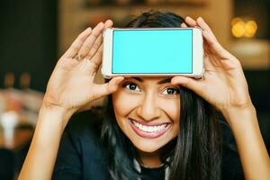 ein lächelnd jung indisch Frau posieren mit ihr Telefon gehaltenen zu ihr Stirn foto