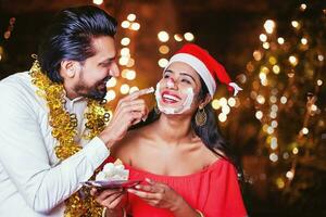 glücklich indisch Mann bewirbt sich Kuchen Sahne auf Frau Gesicht auf Weihnachten Nacht foto