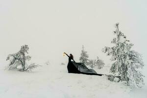 Fee schön Frau im ein lange schwarz Kleid hält ein alt Laterne im ihr Hand im ein schneebedeckt Wald gegen das Hintergrund von ein nebelig Winter Hügel und Schneefall. schwarz Hexe kommt aus von das schneebedeckt Wald. foto