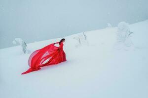 Fee Geschichte Mädchen auf Polar- Winter Landschaft. jung schön Frau im lange rot Kleid Über Winter Hügel Hintergrund und Schneefall. foto
