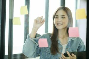 jung weiblich Schüler Schreiben Studie Pläne auf Post-its auf Glas Mauer. foto