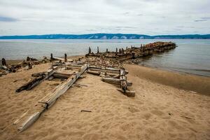 alt gebrochen Boot Seebrücke auf das Insel von olchon auf See Baikalsee. verfault Riegel und Protokolle. auf das Ufer von Steine und Sand. hinter das Berg See. das Wetter ist düster, das Himmel ist grau. foto