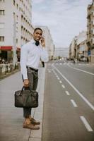 junger afroamerikanischer Geschäftsmann, der ein Mobiltelefon benutzt, während auf ein Taxi auf einer Straße wartet foto