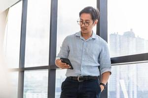 asiatischer Geschäftsmann, der neben großem Fenster steht und Handy betrachtet foto