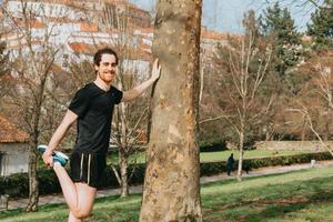 Ein junger Mann streckte sein Bein gegen einen Baum, während er im Park mit Kopierraum trainierte foto
