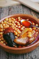 Kichererbsenwurst und Speck in einem Topf mit Holzlöffel und Gabel und einem kleinen Metalllöffel foto