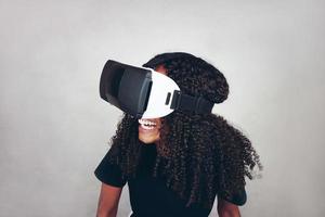 Eine schöne junge schwarze Frau mit lockigem Afro-Haar trägt Virtual Reality VR Headset und spielt Videospiele, während sie im Studio mit grauem Hintergrund lächelt foto