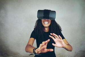 Eine schöne junge schwarze Frau mit lockigem Afro-Haar trägt Virtual Reality VR Headset und spielt Videospiele, während sie im Studio mit grauem Hintergrund lächelt foto