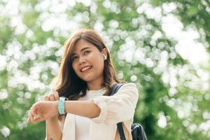 glücklich jung asiatisch Frau Gehen im das Park und mit Clever Uhr foto
