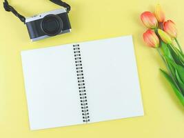 eben legen von leer Seite geöffnet Notizbuch, Digital Kamera und rot Gelb Tulpen Blumen auf Gelb Hintergrund mit Kopieren Raum. foto