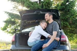 Papa und Sohn sind Sitzung im das Kofferraum von ein Auto beim Sonnenuntergang. umarmen Papa und Lachen foto