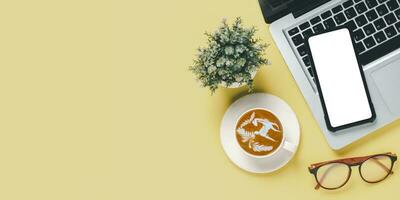 oben Sicht, Gelb Büro Schreibtisch mit Laptop Computer, leer Bildschirm Clever Telefon, Tasse von Kaffee und Brille Kopieren Raum, spotten hoch. foto