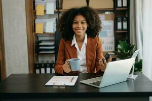 zuversichtlich schön afrikanisch Geschäftsfrau Tippen Laptop Computer und Digital Tablette während halten Kaffee beim Büro foto