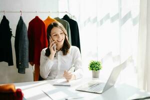 Mode Designer Frau reden Clever Telefon und mit Laptop mit Digital Tablette Computer im modern Studio das Kleider hängend auf das Gestelle foto