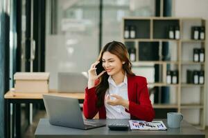 Geschäft asiatisch Frau reden auf das Telefon und mit ein Laptop mit ein Lächeln während Sitzung beim modern Büro foto