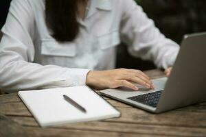 asiatisch jung Frau im Clever beiläufig tragen Schreiben auf Notizbuch und Arbeiten auf Laptop während Sitzung im kreativ Büro oder Cafe. jung Mädchen Arbeiten mit Laptop auf das Holz Tabelle im das Cafe foto