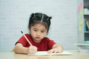 asiatisch Baby Mädchen tragen ein rot Hemd schreiben Anmerkungen im Notizbuch und lesen Buch zu Studie online auf Holz Tabelle Schreibtisch im Leben Zimmer beim heim. Bildung Lernen online von Zuhause Konzept. foto