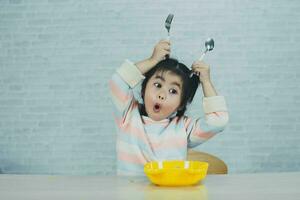 asiatische Babymädchen genießen glücklich mit Bestecklöffel und Gabel, die köstliche Nudeln in der Küche am Esstisch essen. Fröhliches asiatisches Mädchen übt, alleine am Esstisch zu essen. Babynahrungskonzept foto