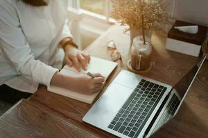 Frau im Clever beiläufig tragen Schreiben auf Notizbuch und Arbeiten auf Laptop während Sitzung im kreativ Büro oder Cafe. jung Mädchen Arbeiten mit Laptop auf das Holz Tabelle im das Cafe foto