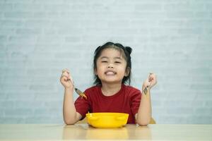 asiatische Babymädchen genießen glücklich mit Bestecklöffel und Gabel, die köstliche Nudeln in der Küche am Esstisch essen. Fröhliches asiatisches Mädchen übt, alleine am Esstisch zu essen. Babynahrungskonzept foto