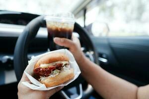 asiatisch Frau Treiber halt und Essen Hamburger im Auto, gefährlich und Risiko ein Unfall. foto