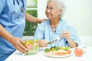 asiatisch Alten Frau geduldig Essen Lachs Anteil und Gemüse Salat zum gesund Essen im Krankenhaus. foto