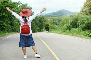 zurück Aussicht von asiatisch Frau Reisender trägt rot Hut und rot Rucksack, erhöht Hände hoch, steht auf das ländlich Straße, Wald Sicht. Konzept, Reisen allein, Freiheit. Hobby und Erholung Aktivität. foto