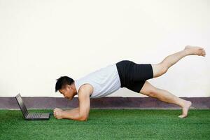 asiatisch Mann ist tun Planke Yoga Übung durch Aufpassen von Computer Laptop. Konzept, Gesundheit Pflege, geben Zeit zu du selbst. Erholung Aktivität. Hobby und gesund Lebensstil. genießen Video foto