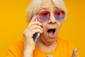 Foto von im Ruhestand alt Dame reden auf das Telefon im Gelb Brille Gelb Hintergrund