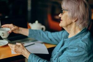 schön reifen Senior Frau mit Brille sitzt beim ein Tabelle im Vorderseite von ein Laptop Sozial Netzwerke unverändert foto