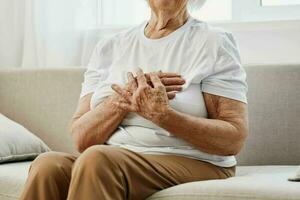 Alten Frau stark Truhe Schmerzen Sitzung auf das Sofa, Gesundheit Probleme im alt Alter, Arm Qualität von Leben. Oma mit grau Haar hält ihr Truhe mit ihr Hände, Damen Gesundheit, Brust Krebs. foto