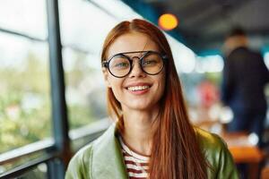 Frau Hipster mit rot Haar und Brille sitzt im Stadt, Dorf im Cafe Lächeln mit Zähne suchen beim Kamera, Frau freiberuflich Blogger Nahansicht foto