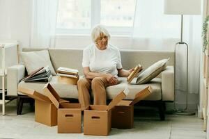 Alten Frau sitzt auf ein Sofa beim Zuhause mit Boxen. Sammeln Dinge mit Erinnerungen Alben mit Fotos und Foto Frames ziehen um zu ein Neu Platz Reinigung Dinge und ein glücklich lächeln. Lebensstil Ruhestand.