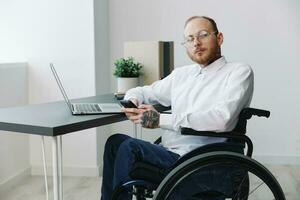 ein Mann Rollstuhl im das Büro funktioniert beim ein Laptop mit ein Telefon, Arbeiten online, Sozial Netzwerke und Start-up, Integration in Gesellschaft, das Konzept Arbeiten Person mit Behinderungen, echt Person Nahansicht foto