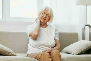 Alten Frau stark Hals Schmerzen Sitzung auf das Sofa, Gesundheit Probleme im alt Alter, Arm Qualität von Leben. foto