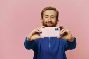 Porträt von ein rothaarig Mann mit Telefon im Hand nehmen Selfies und Fotos auf seine Telefon mit ein Lächeln auf ein Rosa Hintergrund, Blogger