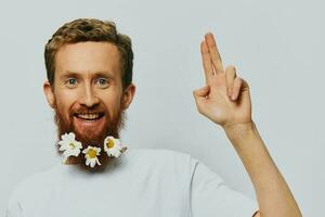 Porträt von ein komisch Mann im ein Weiß T-Shirt mit Blumen Gänseblümchen im seine Bart auf ein Weiß isoliert Hintergrund, Kopieren Ort. Urlaub Konzept und Glückwunsch. foto