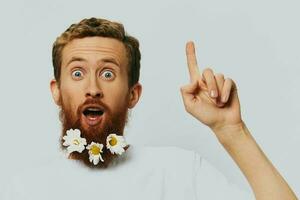 Porträt von ein komisch Mann im ein Weiß T-Shirt mit Blumen Gänseblümchen im seine Bart auf ein Weiß isoliert Hintergrund, Kopieren Ort. Urlaub Konzept und Glückwunsch. foto