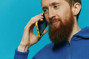 Porträt von ein Mann mit ein Telefon im seine Hände tut suchen beim es und reden auf das Telefon, auf ein Blau Hintergrund. kommunizieren online Sozial Medien, Lebensstil foto