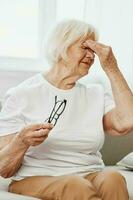 Alten Frau stark Auge Schmerzen Sitzung auf das Couch, Gesundheit Probleme im alt Alter, Arm Qualität von Leben. ein Oma mit grau Haar hält auf zu ihr wund Augen mit Brille im ihr Hand. foto