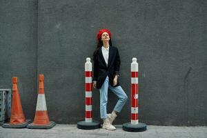 Mode Frau Stehen gelehnt gegen ein Mauer Straße gegen ein Hintergrund von das Stadt Straße funktioniert Tourist im stilvoll Kleider mit rot Lippen und rot Baskenmütze, reisen, filmisch Farbe, retro Jahrgang Stil. foto