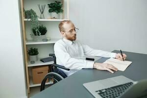 ein Mann Rollstuhl Geschäftsmann im das Büro funktioniert beim ein Laptop, Schreiben Nieder Ideen und ein planen im ein Notizbuch, Nachdenklichkeit, Integration in Gesellschaft, das Konzept von Arbeiten ein Person mit Behinderungen foto
