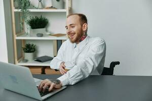 ein Mann im ein Rollstuhl ein Geschäftsmann mit Tätowierungen im das Büro funktioniert beim ein Laptop mit Kaffee, Integration in Gesellschaft, das Konzept von Arbeiten ein Person mit Behinderungen, Freiheit von Sozial Rahmen foto