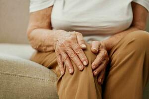 Alten Frau stark Schmerzen im ihr Bein Sitzung auf das Couch, Gesundheit Probleme im alt Alter, Arm Qualität von Leben. Oma mit grau Haar hält auf zu ihr wund Knie, Probleme mit Gelenke und Bänder. foto