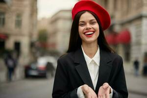 Mode Frau Porträt Lächeln Zähne Stehen auf das Straße im das Stadt Hintergrund im stilvoll Kleider mit rot Lippen und rot Baskenmütze, reisen, filmisch Farbe, retro Jahrgang Stil, städtisch Mode Lebensstil. foto