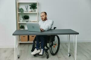 ein Mann im ein Rollstuhl ein Geschäftsmann mit Tätowierungen im das Büro funktioniert beim ein Laptop, Integration in Gesellschaft, das Konzept von Arbeiten ein Person mit Behinderungen, Freiheit von Sozial Frameworks foto
