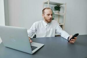 ein Mann im ein Rollstuhl ein Geschäftsmann im das Büro funktioniert beim ein Laptop und hält ein Telefon im seine Hände, Nachdenklichkeit, Integration in Gesellschaft, das Konzept von Arbeiten ein Person mit Behinderungen foto