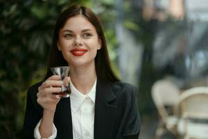 Geschäft Brünette Frau mit rot Lippen Lächeln mit Zähne Getränke Tee im ein Cafe von ein Türkisch Glas Becher im ein Weiß Hemd und schwarz Mode Jacke auf das Straße, Sommer- reisen, Ferien im das Stadt foto