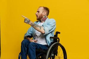 ein Mann im ein Rollstuhl Probleme mit das Bewegungsapparat System sieht aus beim das Kamera zeigt an ein Finger An, mit Tätowierungen auf seine Hände sitzt auf ein Gelb Studio Hintergrund, Gesundheit Konzept Mann mit Behinderungen foto