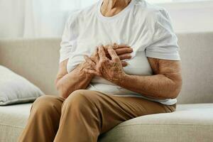 Alten Frau stark Truhe Schmerzen Sitzung auf das Sofa, Gesundheit Probleme im alt Alter, Arm Qualität von Leben. Oma mit grau Haar hält ihr Truhe mit ihr Hände, Damen Gesundheit, Brust Krebs. foto